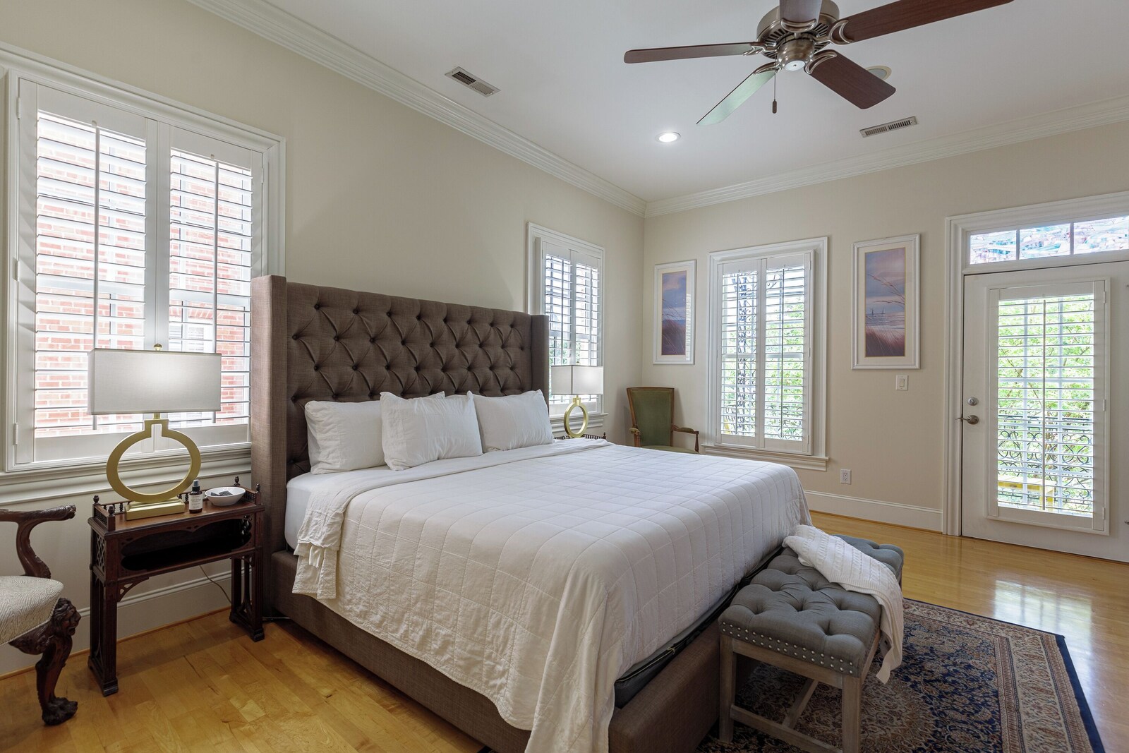 Primary bedroom from the side: tufted king bed, ceiling fan, hardwood floors and a glass door with shutters opening to a balcony