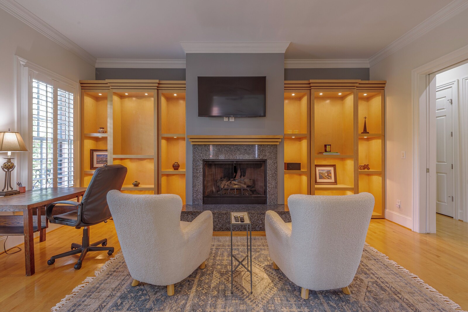 Upstairs lounge at John's Downtown Greensboro townhome: a fireplace with a wall-mounted TV, lit built-in maple bookshelves on both sides, two cream boucle armchairs on a patterned rug, and a desk by a shuttered window