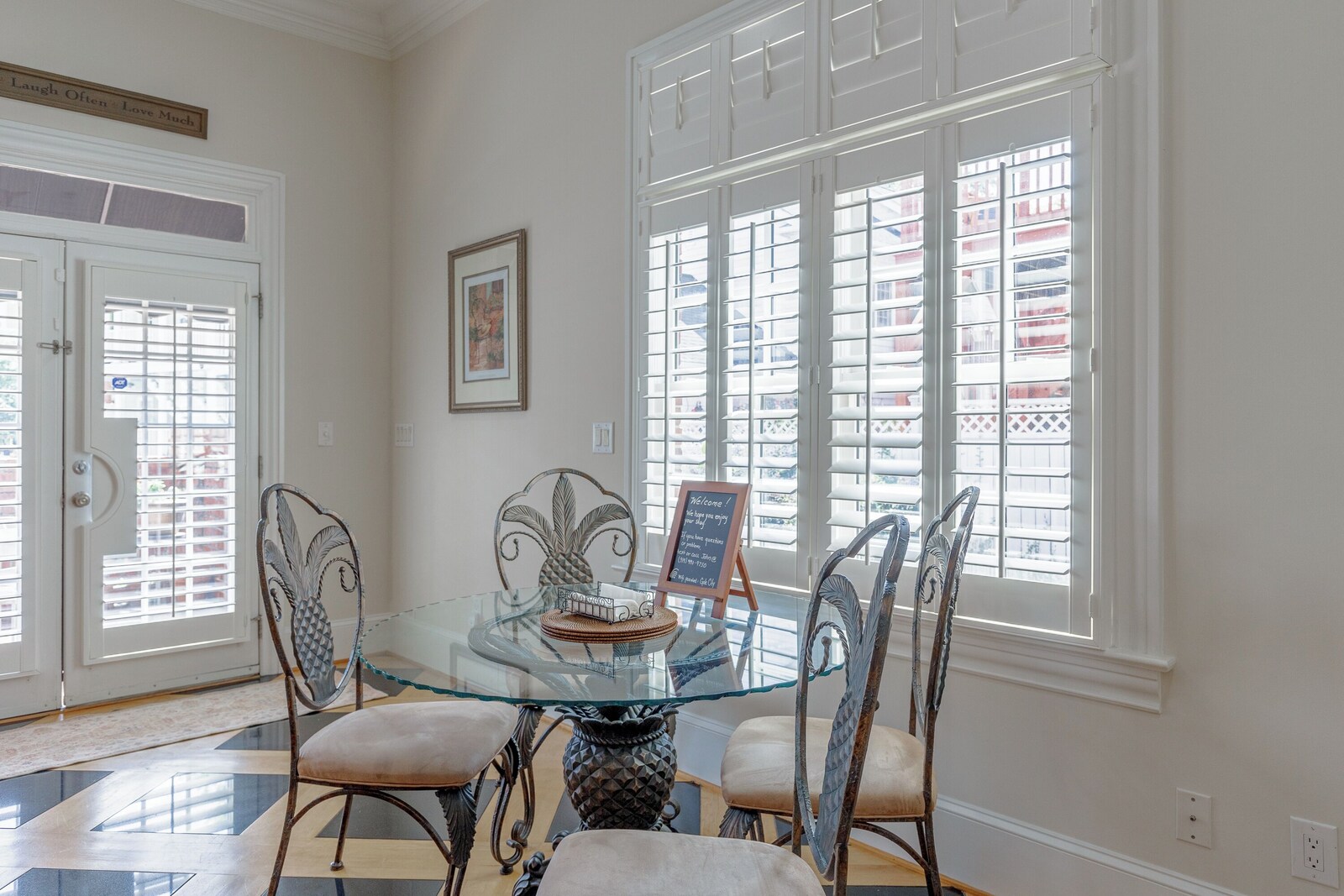 Breakfast nook: round glass table with four wrought-iron pineapple-back chairs and a welcome chalkboard, beside tall shuttered windows and the front door